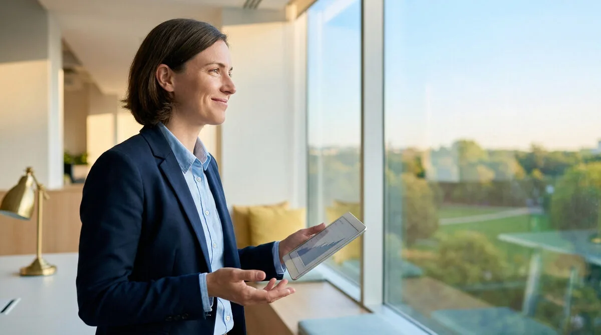 Femme d'affaires souriante en costume bleu, tenant une tablette avec un graphique financier, regarde à travers une fenêtre. Bureau lumineux.